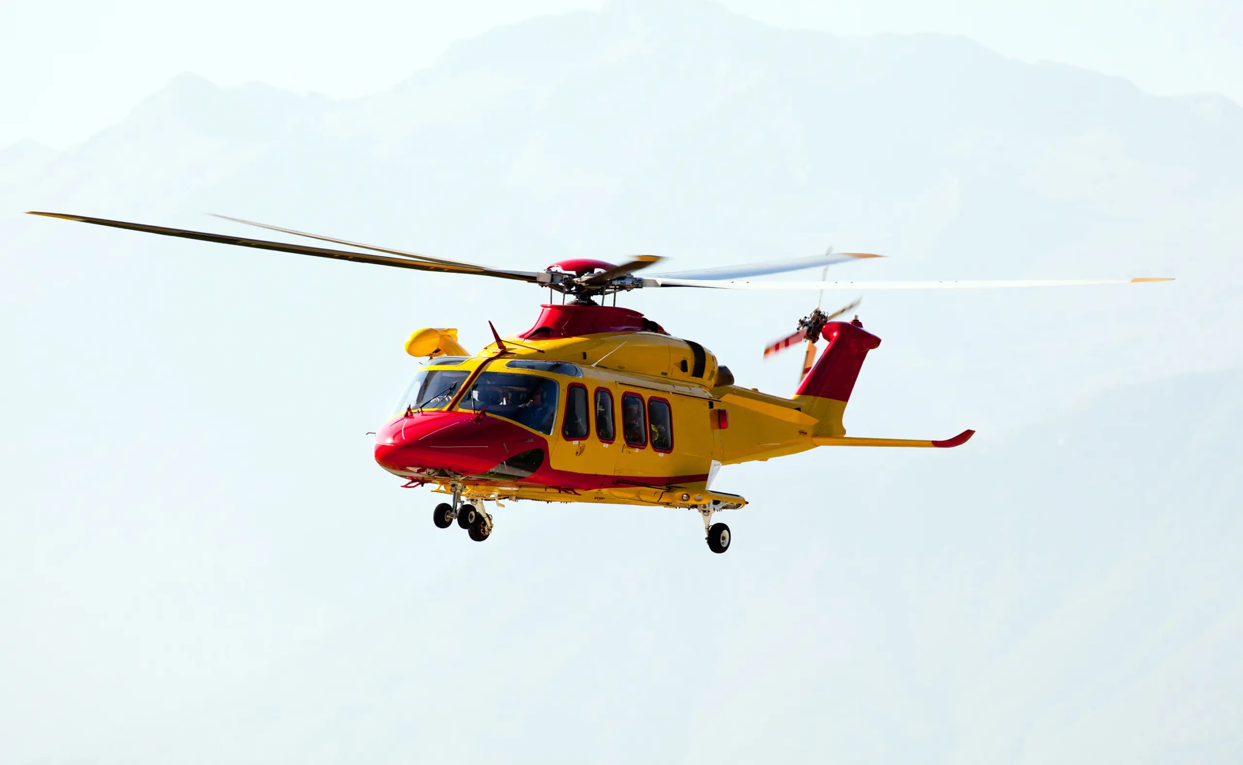 Yellow and red helicopter in flight against a mountainous backdrop.