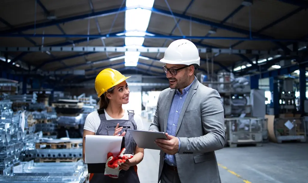 Engineer and worker discussing project details in a factory, both wearing safety helmets.