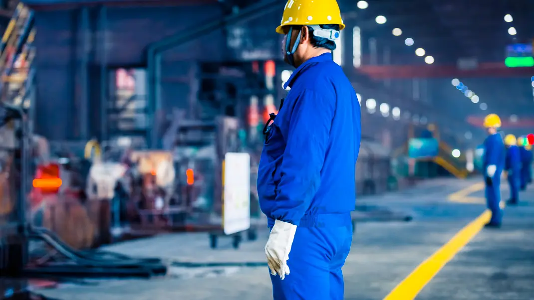 Factory worker in blue overalls and yellow helmet observing machinery in an industrial setting.
