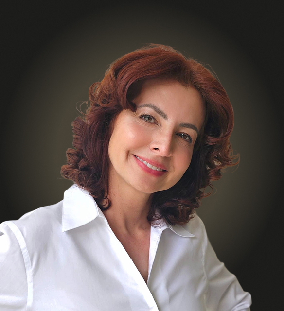 Smiling woman with curly hair wearing a white blouse against a dark background.