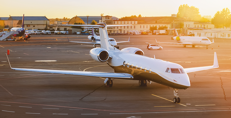 Private jet on a sunny tarmac at an airport, surrounded by other small aircraft and hangars in the background.
