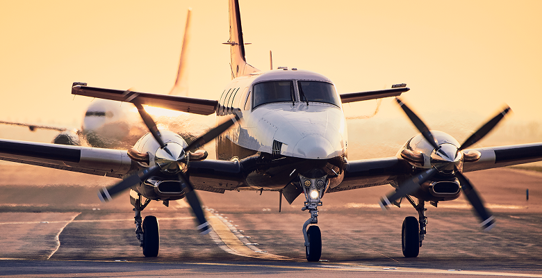 Twin-engine propeller plane taxiing on runway at sunrise, with blurred plane in background.